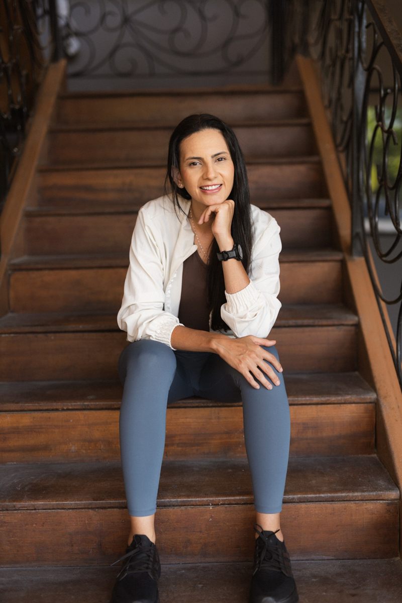 Kamila Rodrigues sentada numa escada de madeira, de jaqueta branca, sorrindo